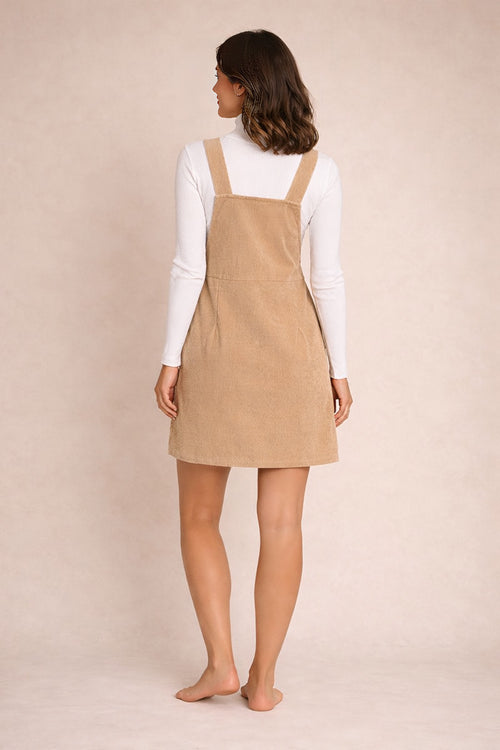 Woman wearing a beige pinafore dress over a white shirt against a beige background