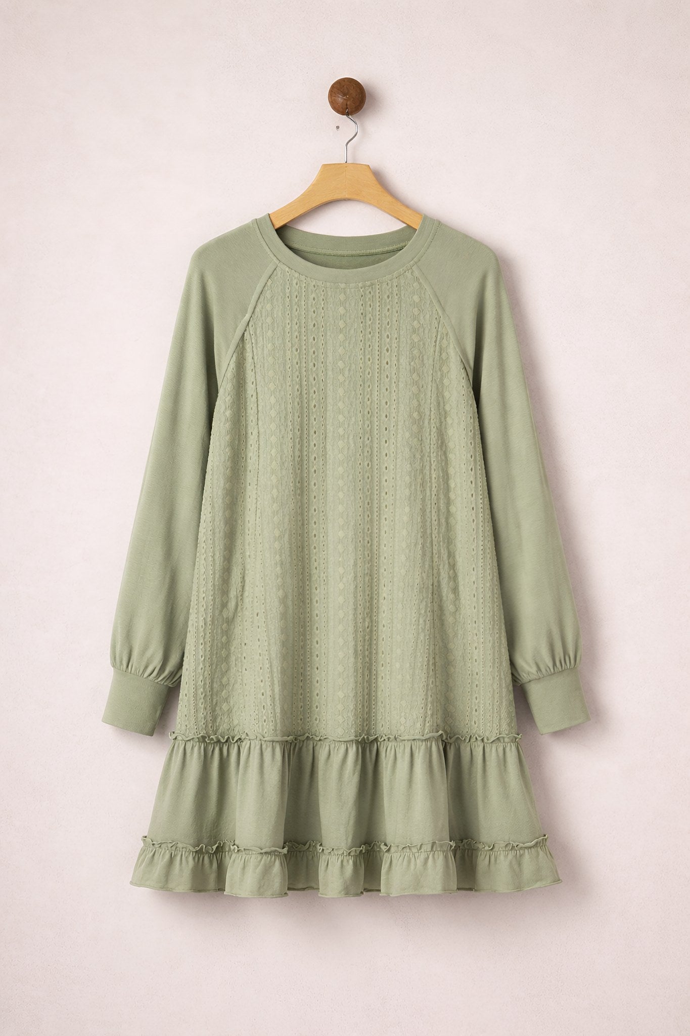 Green dress with long sleeves and ruffled hem on a wooden hanger against a light pink background